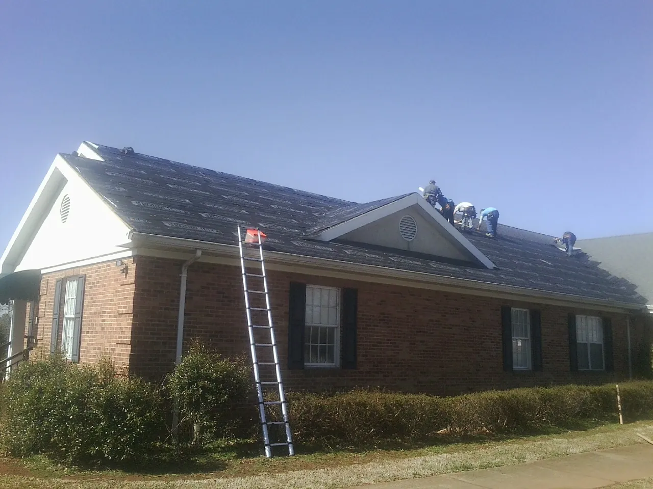 Crew tearing shingles off a brick ranch roof with a ladder set — Emergency Roof Repair work by our crew in Mead